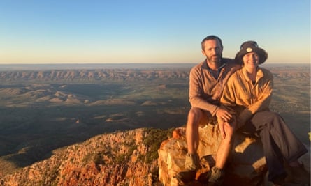 Tess and Piers at Mount Sonder, Northern Territory in 2022. They are seated on a rock lit up by the golden light of a setting sun shining from the left of the photo, while behind them can be seen an expansive green valley with a line of mountainous terrain near the horizon.