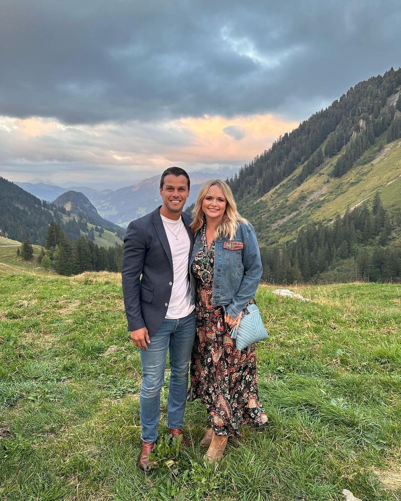 Miranda and Brendan stand on a grassy hilltop surrounded by mountains
