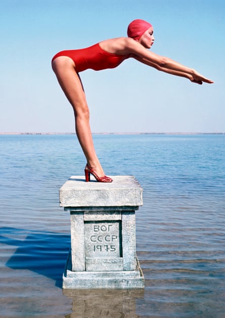 American model Jerry Hall on a plinth in the sea in the USSR, wearing a red swimsuit,  swimming cap and sandals, looking as if she’s about to dive in, 1975