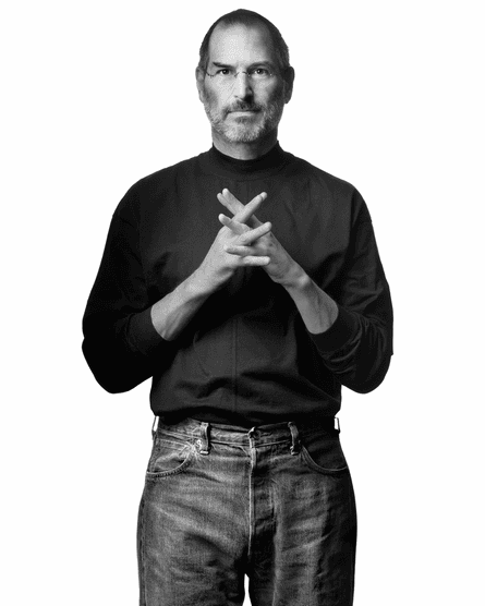 Steve Jobs, fingers locked in front of him, wearing black roll-neck and jeans, against white background